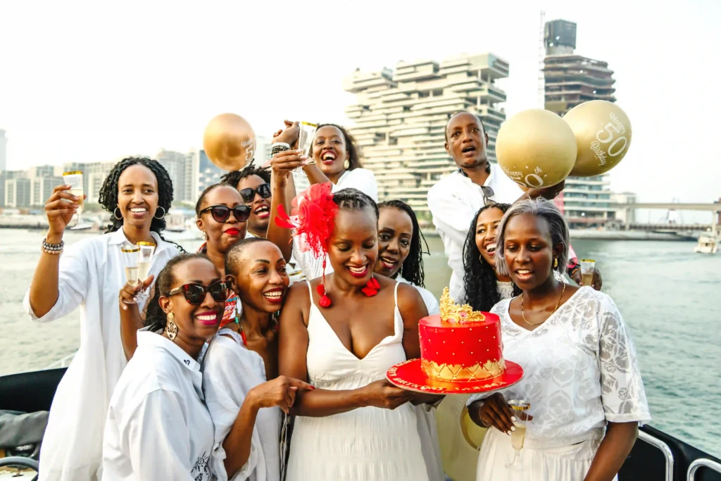 A joyful 50th birthday party celebration on a boat, captured by professional birthday photographer in Dubai. Guests dressed in white smile around a woman holding a red cake with a modern city waterfront backdrop.